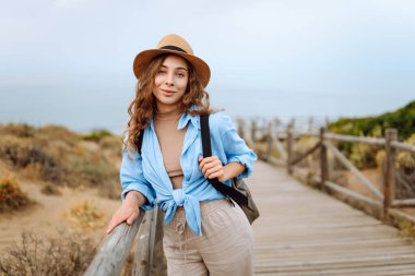 Hasır şapkalı ve sırt çantalı güzel bir turist deniz boyunca tahta bir patika boyunca yürüyor. Genç bir kadın doğayı seviyor, güzel manzarayı. Seyahat, yürüyüş, tatil konsepti.