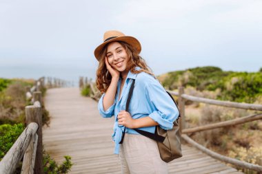 Hasır şapkalı ve sırt çantalı güzel bir turist deniz boyunca tahta bir patika boyunca yürüyor. Genç bir kadın doğayı seviyor, güzel manzarayı. Seyahat, yürüyüş, tatil konsepti.