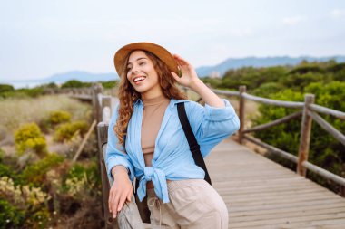 Hasır şapkalı ve sırt çantalı güzel bir turist deniz boyunca tahta bir patika boyunca yürüyor. Genç bir kadın doğayı seviyor, güzel manzarayı. Seyahat, yürüyüş, tatil konsepti.