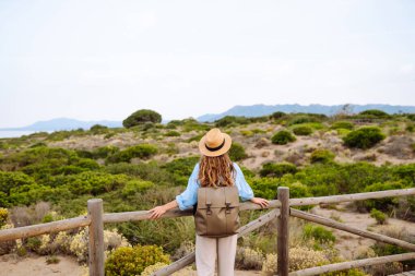 Hasır şapkalı ve sırt çantalı güzel bir turist deniz boyunca tahta bir patika boyunca yürüyor. Genç bir kadın doğayı seviyor, güzel manzarayı. Seyahat, yürüyüş, tatil konsepti.