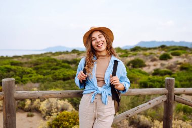 Hasır şapkalı ve sırt çantalı güzel bir turist deniz boyunca tahta bir patika boyunca yürüyor. Genç bir kadın doğayı seviyor, güzel manzarayı. Seyahat, yürüyüş, tatil konsepti.