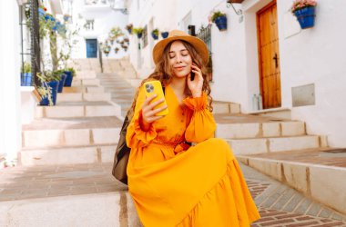 Güzel bir kadın eski Avrupa şehrinde telefonla yürüyor. Sırt çantalı ve parlak elbiseli genç turist güneşli havanın tadını çıkarıyor. İletişim, teknoloji, seyahat kavramı.