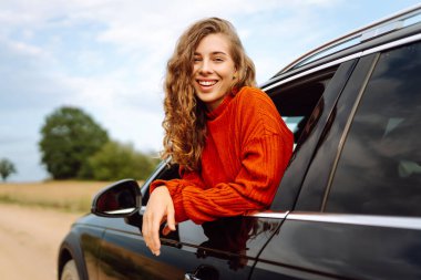 Arabanın camından sarkan, özgürlüğü hisseden, güneşli günün tadını çıkaran genç bir kadın. Şık giyinmiş güzel bir kadın, araba sürerken eğleniyor. Seyahat konsepti, özgürlük, araba maceraları.