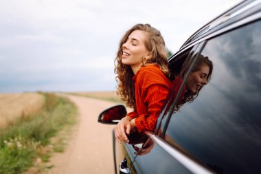 Arabanın camından sarkan, özgürlüğü hisseden, güneşli günün tadını çıkaran genç bir kadın. Şık giyinmiş güzel bir kadın, araba sürerken eğleniyor. Seyahat konsepti, özgürlük, araba maceraları.