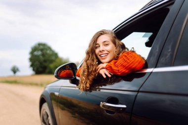 Arabanın camından sarkan, özgürlüğü hisseden, güneşli günün tadını çıkaran genç bir kadın. Şık giyinmiş güzel bir kadın, araba sürerken eğleniyor. Seyahat konsepti, özgürlük, araba maceraları.