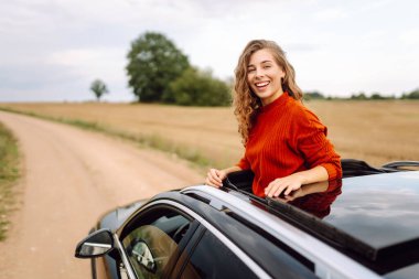 Arabanın camından sarkan, özgürlüğü hisseden, güneşli günün tadını çıkaran genç bir kadın. Şık giyinmiş güzel bir kadın, araba sürerken eğleniyor. Seyahat konsepti, özgürlük, araba maceraları.