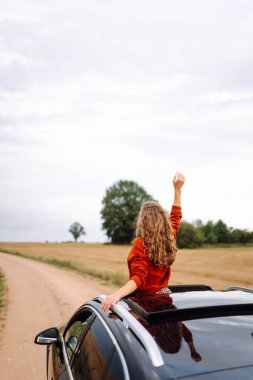 Arabanın camından sarkan, özgürlüğü hisseden, güneşli günün tadını çıkaran genç bir kadın. Şık giyinmiş güzel bir kadın, araba sürerken eğleniyor. Seyahat konsepti, özgürlük, araba maceraları.