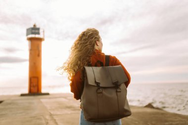 Sırt çantalı hoş bir kadın deniz fenerinde günbatımının tadını çıkarıyor, özgürlüğü hissediyor. Turist doğada dinleniyor, sette yürüyor. Yürüme, seyahat, özgürlük kavramı. Yaşam biçimi.