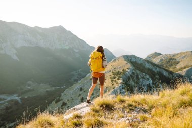 Genç bayan turist dağın tepesinde duruyor, uçurumda günbatımının tadını çıkarıyor. Sırt çantalı gezgin özgürlüğü hisseder, dağ manzaralarına bakar. Seyahat konsepti, özgürlük. Etkin yaşam biçimi.