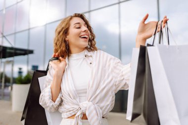 Şık elbiseli ve kıvırcık saçlı genç bir kadın alışveriş merkezinin yakınında elinde alışveriş torbalarıyla yürüyor. Mutlu kadın güneşli günün tadını çıkarıyor. Alışveriş konsepti, satış. Yaşam biçimi.