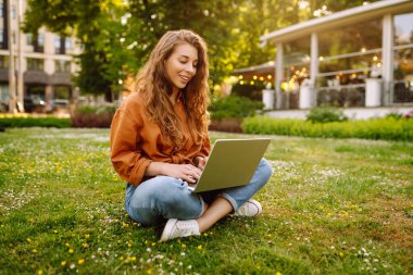 Yeşil çimenlikteki güneşli bir parkta dizüstü bilgisayarı olan mutlu bir kadın. Genç bayan serbest çalışan dışarıda havanın tadını çıkarıyor. Uzaktan çalışma konsepti. Etkin yaşam biçimi.