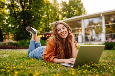 Yeşil çimenlikteki güneşli bir parkta dizüstü bilgisayarı olan mutlu bir kadın. Genç bayan serbest çalışan dışarıda havanın tadını çıkarıyor. Uzaktan çalışma konsepti. Etkin yaşam biçimi.
