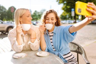 Kafedeki bir masada selfie çeken iki genç arkadaş. Mutlu kadınlar açık havada telefonla vakit geçiriyorlar. Selfie zamanı. Yürüme, blog yazma kavramı.