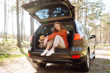 Şık giyinmiş genç bir kadın bir arabanın bagajında dinleniyor. Güneşli havanın tadını çıkaran güzel bir kadın. Araba konseptiyle seyahat etmek. Etkin yaşam biçimi.