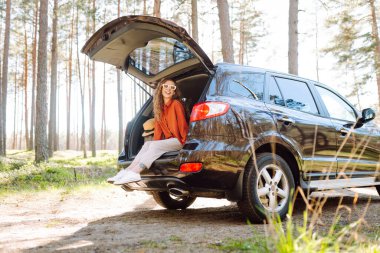 Şık giyinmiş genç bir kadın bir arabanın bagajında dinleniyor. Güneşli havanın tadını çıkaran güzel bir kadın. Araba konseptiyle seyahat etmek. Etkin yaşam biçimi.