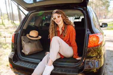 Şık giyinmiş genç bir kadın bir arabanın bagajında dinleniyor. Güneşli havanın tadını çıkaran güzel bir kadın. Araba konseptiyle seyahat etmek. Etkin yaşam biçimi.