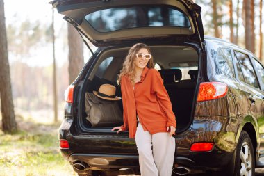 Şık giyinmiş genç bir kadın bir arabanın bagajında dinleniyor. Güneşli havanın tadını çıkaran güzel bir kadın. Araba konseptiyle seyahat etmek. Etkin yaşam biçimi.