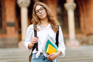 Kıvırcık saçlı güzel bir öğrenci bir üniversite binasının arka planında bir yığın kitapla ayakta duruyor. Elinde kağıtları olan gözlüklü genç bir kadın. Eğitim kavramı, yürüyüşler.