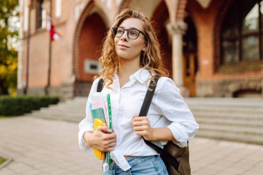 Kıvırcık saçlı güzel bir öğrenci bir üniversite binasının arka planında bir yığın kitapla ayakta duruyor. Elinde kağıtları olan gözlüklü genç bir kadın. Eğitim kavramı, yürüyüşler.