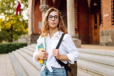 Kıvırcık saçlı güzel bir öğrenci bir üniversite binasının arka planında bir yığın kitapla ayakta duruyor. Elinde kağıtları olan gözlüklü genç bir kadın. Eğitim kavramı, yürüyüşler.
