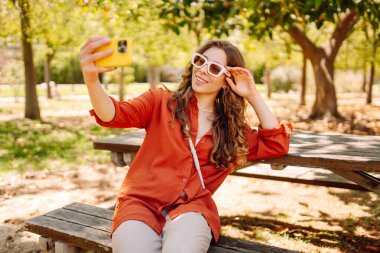 Şık giyinmiş genç bir kadın parkta bir masada otururken selfie çekiyor. Güzel turist dışarıda blog yazıyor. Turizm, blog yazma ve keyif konsepti.