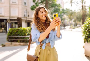Elinde telefon olan güzel bir turist güneşli bir parkta havanın tadını çıkarıyor. Şık giyinmiş genç bir kadın telefonuyla yürüyor. Günlük konsepti.