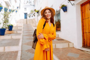 Şapkalı ve parlak elbiseli hoş bir kadın şehrin parlak caddelerinde yürüyor. Sırt çantalı güzel bir kadın yolculuğun tadını çıkarıyor. Turizm, yürüyüş konsepti. Etkin yaşam biçimi.