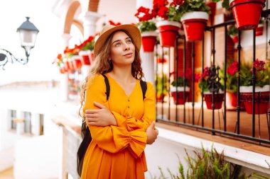 Şapkalı ve parlak elbiseli hoş bir kadın şehrin parlak caddelerinde yürüyor. Sırt çantalı güzel bir kadın yolculuğun tadını çıkarıyor. Turizm, yürüyüş konsepti. Etkin yaşam biçimi.