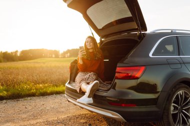 Güzel bir kadın telefonu elinde bir arabanın bagajında gün batımına karşı oturur. Genç bayan dışarıda havanın tadını çıkarıyor. Araba yolculuğu, tatil kavramı. Yaşam biçimi.