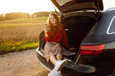 Güzel bir kadın telefonu elinde bir arabanın bagajında gün batımına karşı oturur. Genç bayan dışarıda havanın tadını çıkarıyor. Araba yolculuğu, tatil kavramı. Yaşam biçimi.