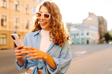 Elinde telefonla şehirde dolaşan neşeli bir kadının portresi. Sokak mimarisinden telefonla keyif alan şık bir kadın turist. Yürüme, keyif alma, blog yazma kavramı.