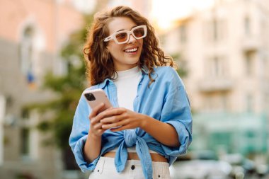 Elinde telefonla şehirde dolaşan neşeli bir kadının portresi. Sokak mimarisinden telefonla keyif alan şık bir kadın turist. Yürüme, keyif alma, blog yazma kavramı.