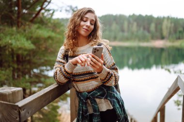 Elinde telefon olan genç bir kadın gölün arka planına karşı. Telefonlu güzel bayan gezgin gün batımının tadını çıkarıyor. Yürüyüş kavramı, özgürlük, keyif.