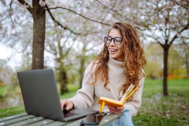 Baharda çiçek açan bir parkta dizüstü bilgisayarı ve defterleri olan genç bir öğrencinin portresi. Dışarıda çalışan güzel bir kadın. Bağımsızlık, eğitim, teknoloji kavramı.