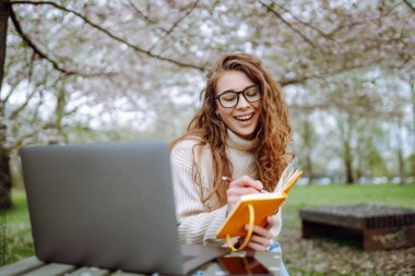 Baharda çiçek açan bir parkta dizüstü bilgisayarı ve defterleri olan genç bir öğrencinin portresi. Dışarıda çalışan güzel bir kadın. Bağımsızlık, eğitim, teknoloji kavramı.