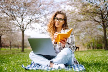 Baharda çiçek açan bir parkta dizüstü bilgisayarı ve defterleri olan genç bir öğrencinin portresi. Dışarıda çalışan güzel bir kadın. Bağımsızlık, eğitim, teknoloji kavramı.
