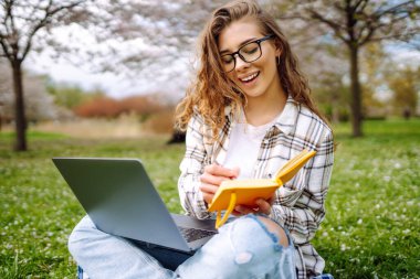 Baharda çiçek açan bir parkta dizüstü bilgisayarı ve defterleri olan genç bir öğrencinin portresi. Dışarıda çalışan güzel bir kadın. Bağımsızlık, eğitim, teknoloji kavramı.