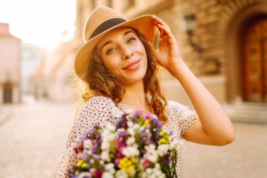 Kıvırcık saçlı, kır çiçekleriyle dolu mutlu bir kadın gün batımının tadını çıkarıyor. Avrupa sokaklarında yürüyen genç turist kadın. Turizm, mutluluk konsepti. Yaşam biçimi.