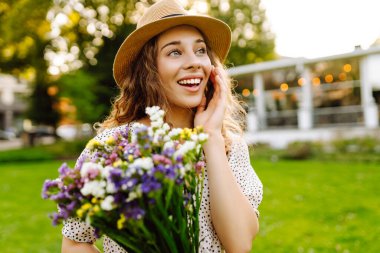 Elinde parlak bir buket olan şapkalı genç bir kadın parkta güneşli bir günün tadını çıkarıyor. Güzel bir kadın dışarıda eğleniyor ve dinleniyor. Rahatlama kavramı, eğlence. Yaşam biçimi.