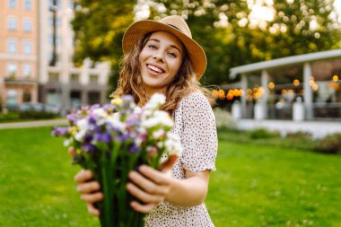 Elinde parlak bir buket olan şapkalı genç bir kadın parkta güneşli bir günün tadını çıkarıyor. Güzel bir kadın dışarıda eğleniyor ve dinleniyor. Rahatlama kavramı, eğlence. Yaşam biçimi.