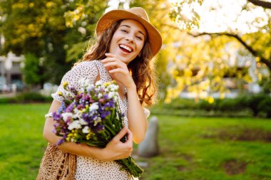 Elinde parlak bir buket olan şapkalı genç bir kadın parkta güneşli bir günün tadını çıkarıyor. Güzel bir kadın dışarıda eğleniyor ve dinleniyor. Rahatlama kavramı, eğlence. Yaşam biçimi.