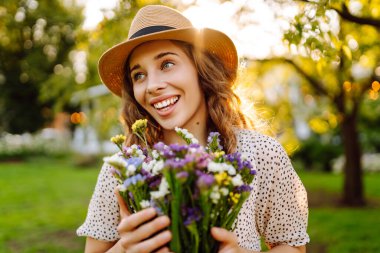 Elinde parlak bir buket olan şapkalı genç bir kadın parkta güneşli bir günün tadını çıkarıyor. Güzel bir kadın dışarıda eğleniyor ve dinleniyor. Rahatlama kavramı, eğlence. Yaşam biçimi.