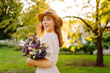 Elinde parlak bir buket olan şapkalı genç bir kadın parkta güneşli bir günün tadını çıkarıyor. Güzel bir kadın dışarıda eğleniyor ve dinleniyor. Rahatlama kavramı, eğlence. Yaşam biçimi.
