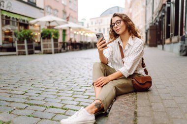 Gözlüklü mutlu kadın elinde telefonla güneşli bir Avrupa caddesinde yürüyor. Genç bir kadın dışarıda telefonuyla eğleniyor. Teknoloji konsepti. Etkin yaşam biçimi.
