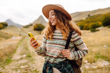 Şapkalı, sırt çantalı ve telefonlu güzel bir kadının portresi. Genç bir kadın telefonda mesajlaşıyor ve dağ manzarasının tadını çıkarıyor. Teknoloji ve seyahat, yürüyüş konsepti.