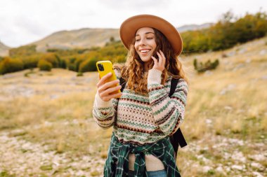Şapkalı, sırt çantalı ve telefonlu güzel bir kadının portresi. Genç bir kadın telefonda mesajlaşıyor ve dağ manzarasının tadını çıkarıyor. Teknoloji ve seyahat, yürüyüş konsepti.