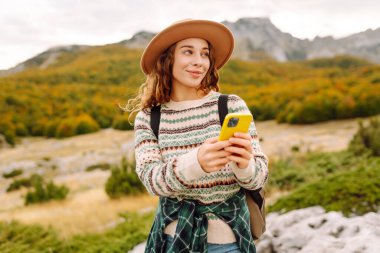 Şapkalı, sırt çantalı ve telefonlu güzel bir kadının portresi. Genç bir kadın telefonda mesajlaşıyor ve dağ manzarasının tadını çıkarıyor. Teknoloji ve seyahat, yürüyüş konsepti.