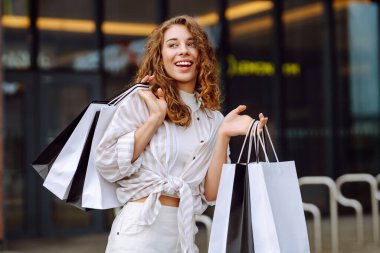 Alışveriş merkezinin yakınında alışveriş çantalı genç bir kadın eğleniyor. Şık giyinmiş bir kadın gün batımında alışveriş çılgınlığında yürüyor. Tüketim, alışveriş kavramı.