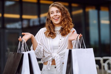 Alışveriş merkezinin yakınında alışveriş çantalı genç bir kadın eğleniyor. Şık giyinmiş bir kadın gün batımında alışveriş çılgınlığında yürüyor. Tüketim, alışveriş kavramı.