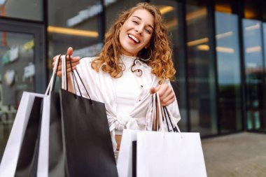 Alışveriş merkezinin yakınında alışveriş çantalı genç bir kadın eğleniyor. Şık giyinmiş bir kadın gün batımında alışveriş çılgınlığında yürüyor. Tüketim, alışveriş kavramı.